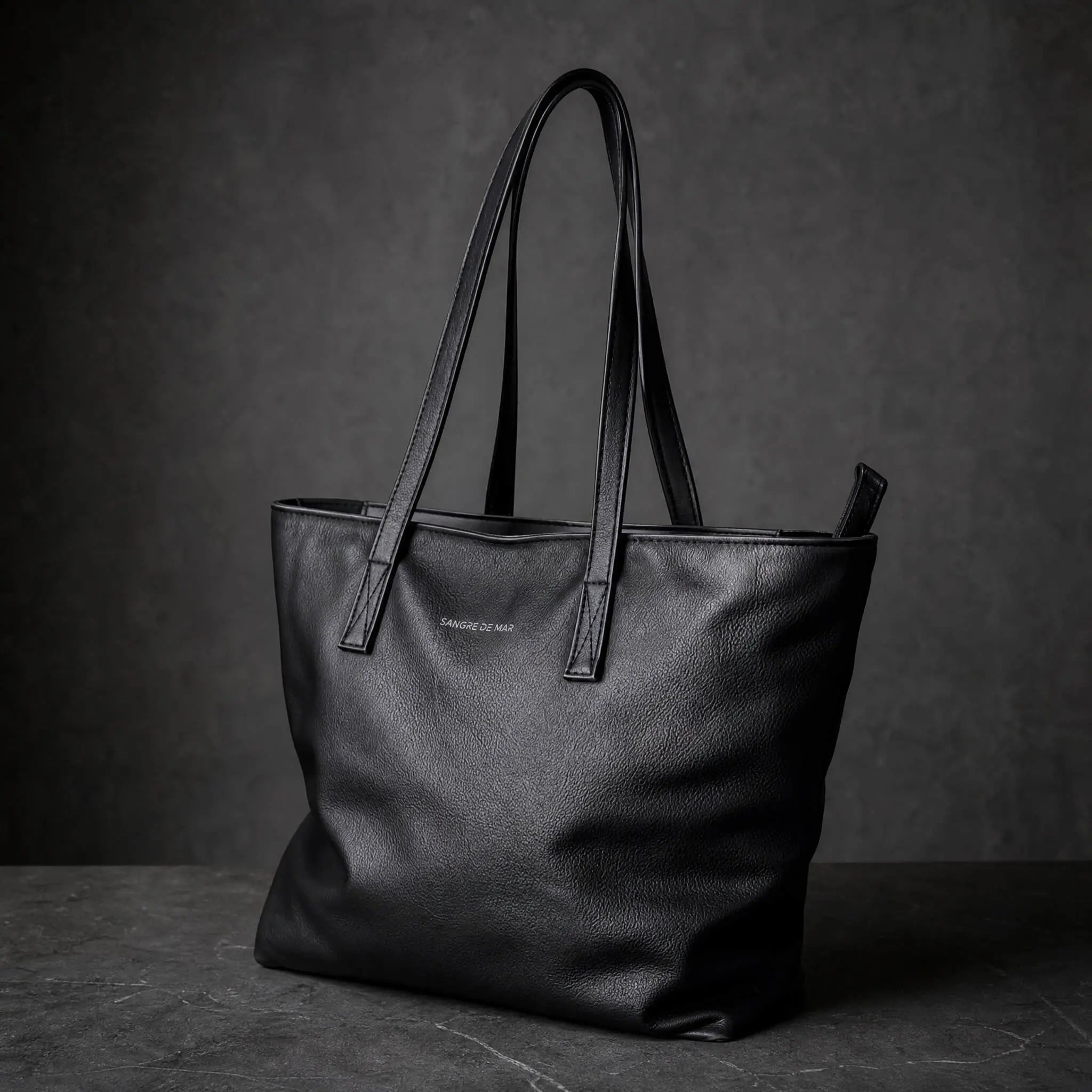 A black pebbled leather tote bag with double shoulder straps, standing on a dark textured surface against a dark gray background. 