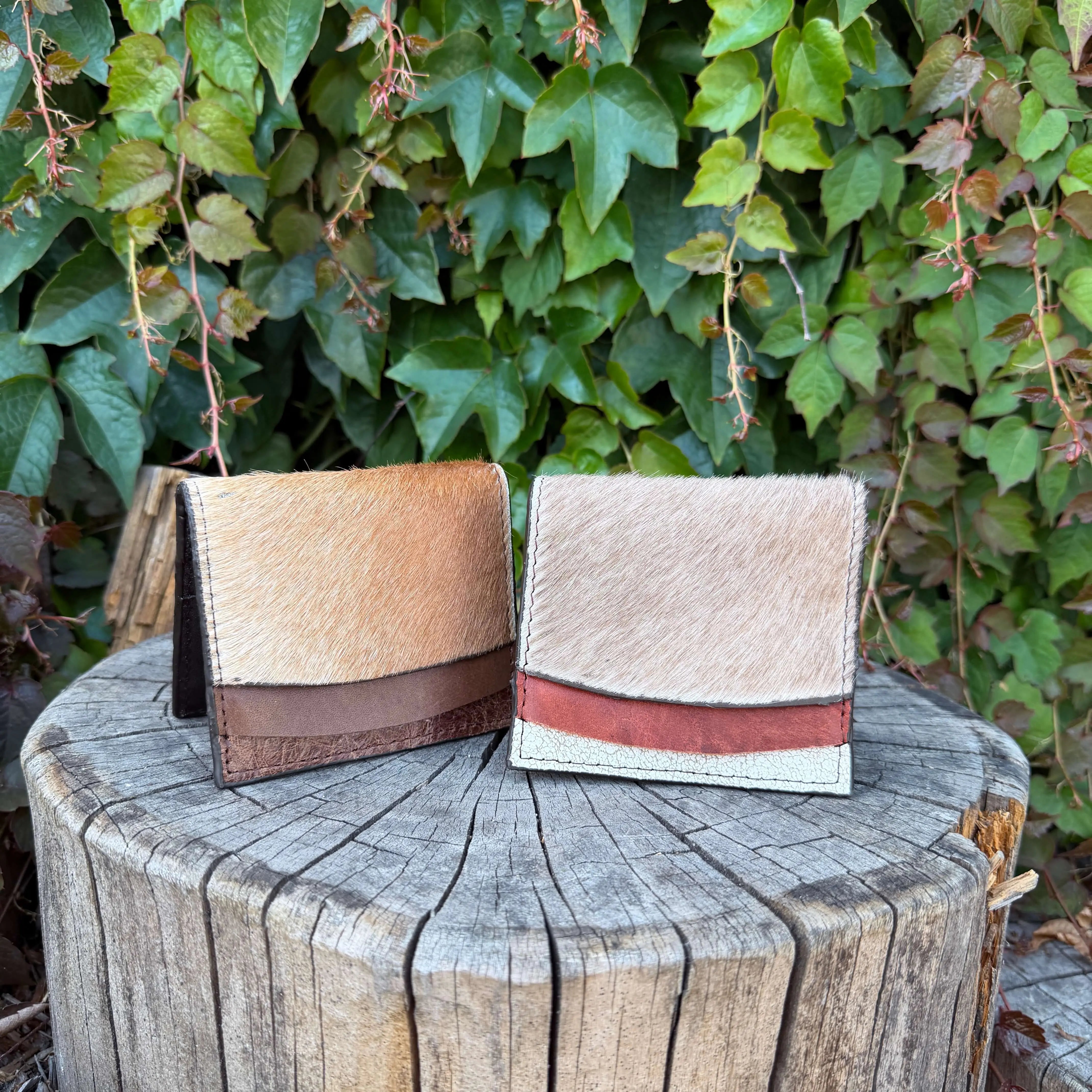 Two cowhide wallets on a wooden stump with green foliage in the background